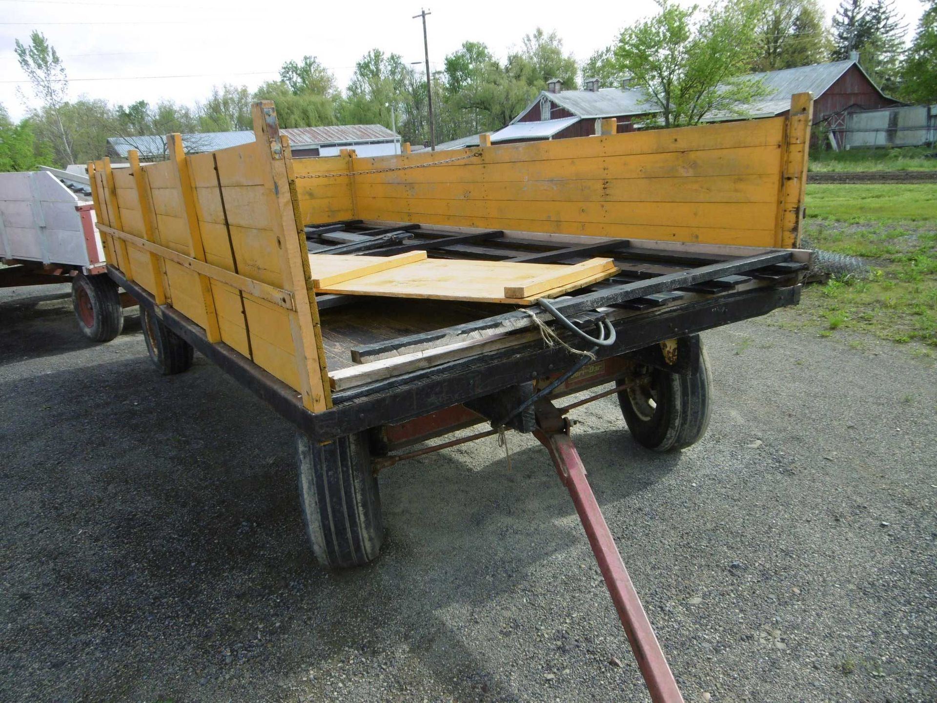 A yellow wooden farm wagon parked on gravel, showing its flatbed and low sides against a backdrop of trees and buildings.