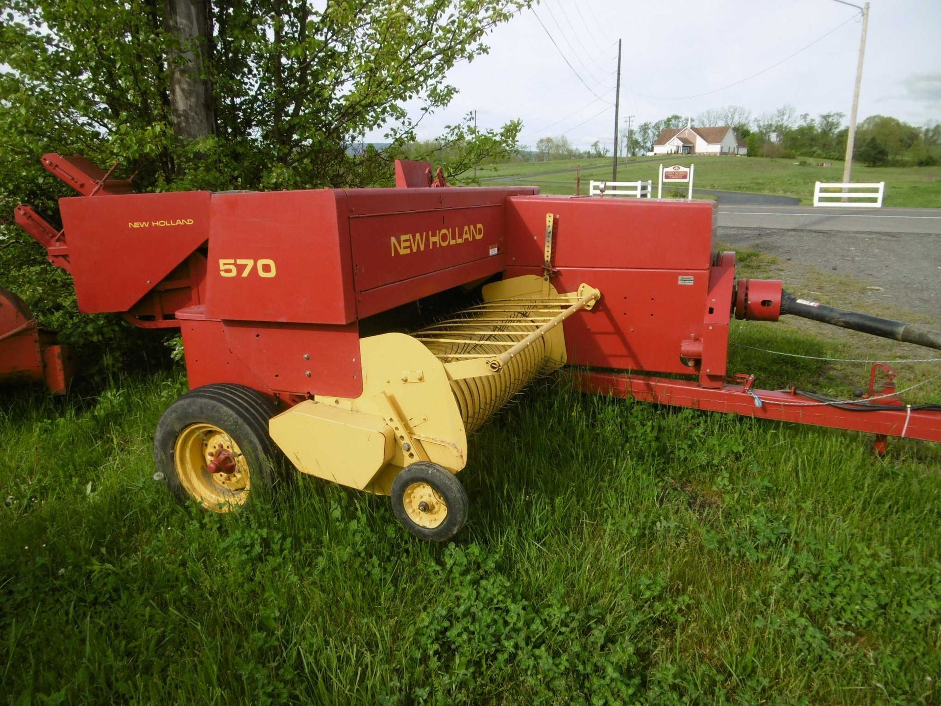 A red New Holland 570 square baler sits in a grassy field on a bright day.
