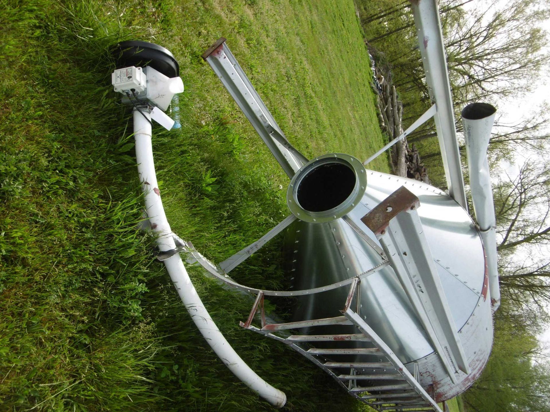 A large, overturned metal grain bin lies on its side in a grassy field with a mechanical auger attached.