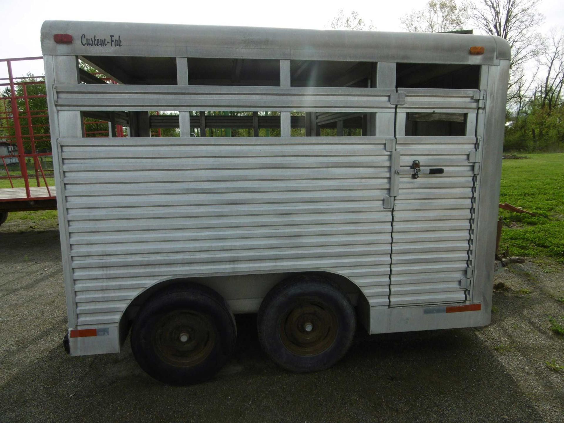 A silver, dual-axle stock trailer for livestock stands on a gravel surface outdoors with green grass in the background.