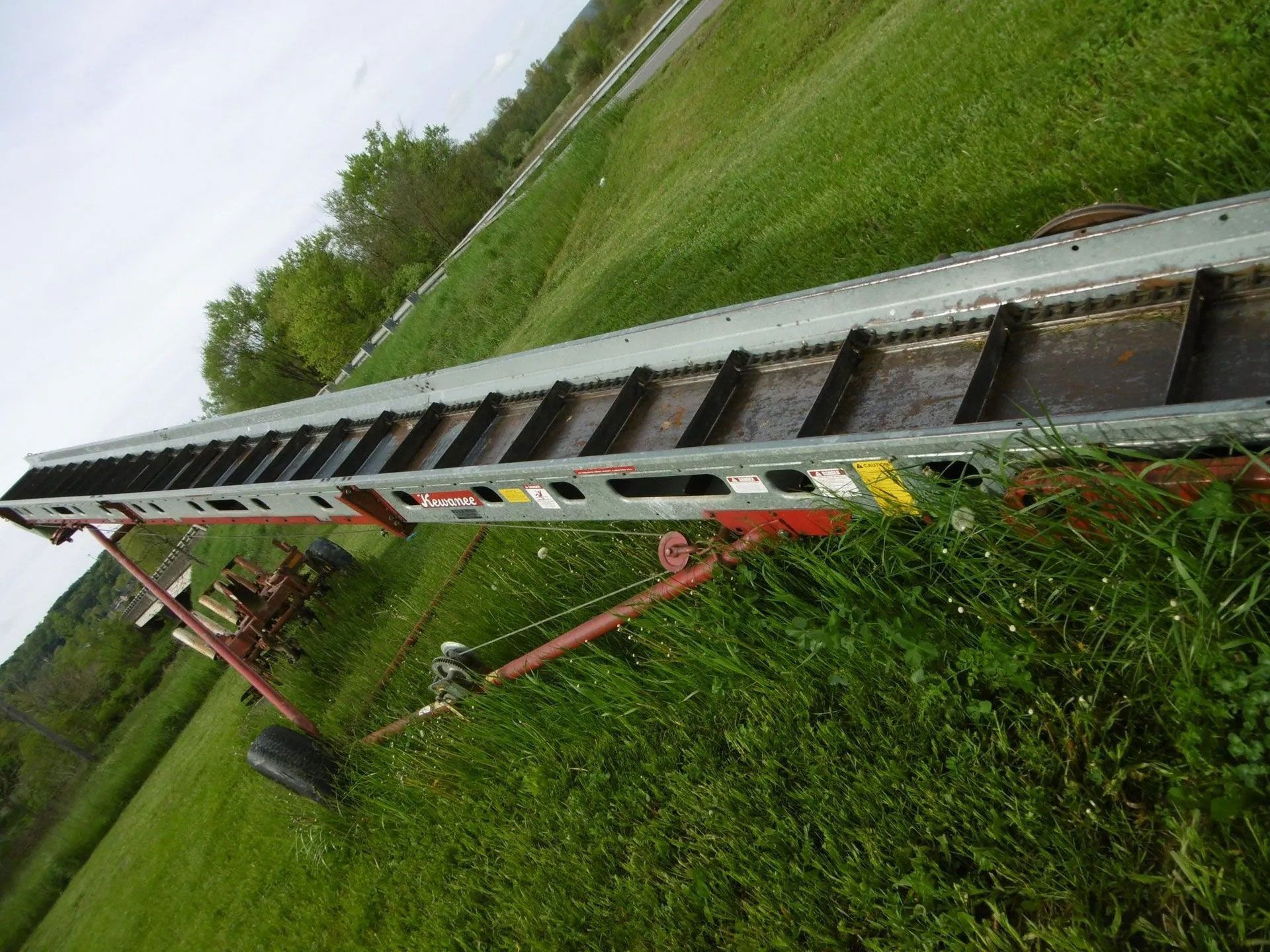 A long, tilted farm conveyor belt with black rungs sits on a grassy field under a cloudy sky.
