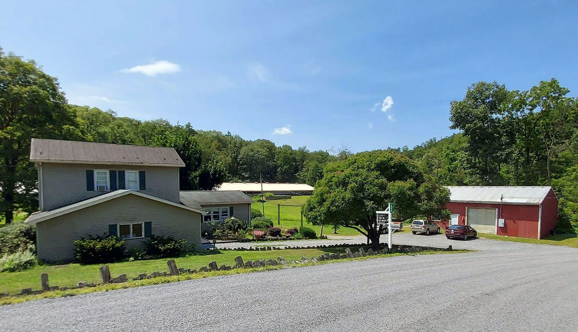 A two-story grey house and a red barn sit on a gravel property surrounded by trees under a bright blue sky.