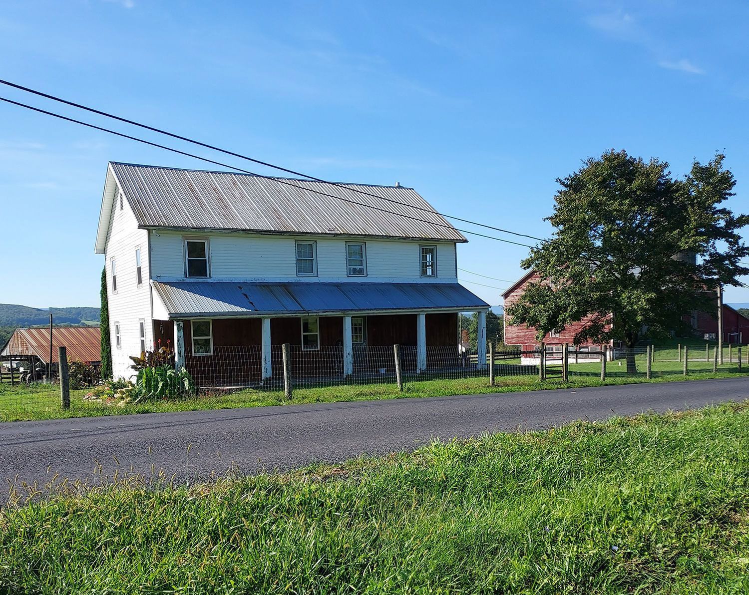 A white, two-story farmhouse with a porch sits beside a road near a red barn and a large tree under a clear blue sky.