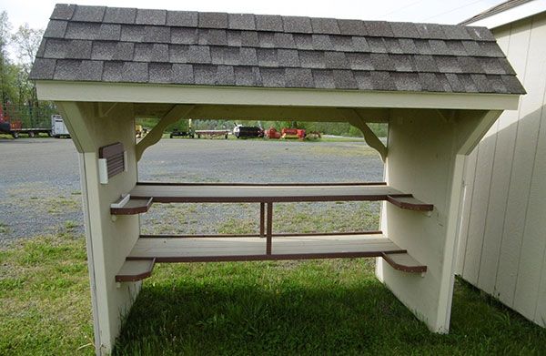 A wooden, roofed outdoor potting bench with two shelves, standing on grass in front of a shed.