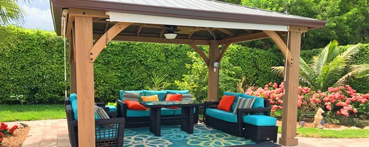 Gazebo with outdoor seating area, blue cushions, table, surrounded by lush greenery.