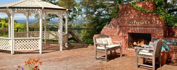 A gazebo and brick fireplace on a patio overlooking a green landscape. Two chairs face the fireplace.
