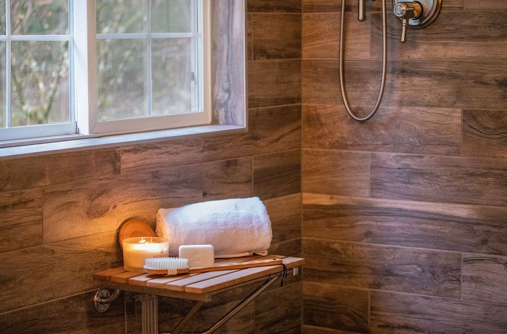 Shower interior with wood-look tile. Shelf holds a candle, soap, and rolled towel next to a window.