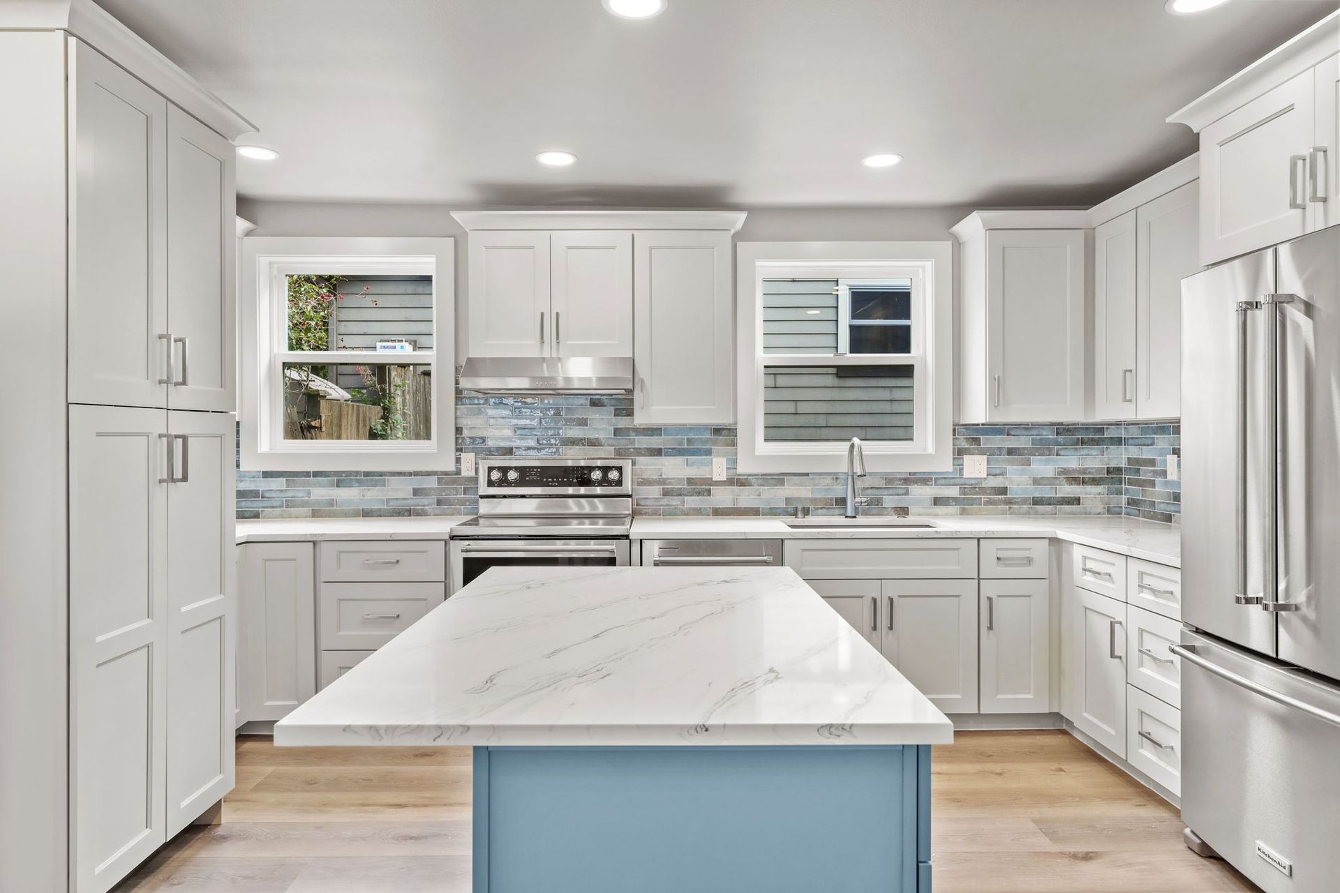 White kitchen with blue backsplash and island, stainless steel appliances, and light wood floors.