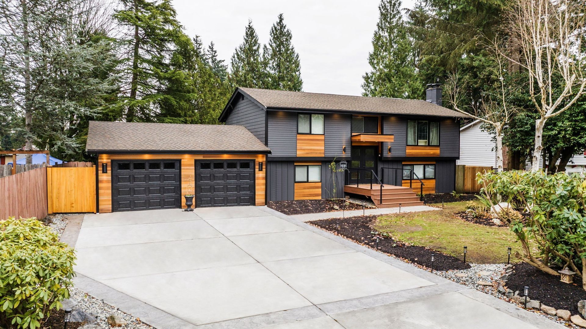 Two-story house with dark gray siding, wood accents, black garage doors, and long driveway.