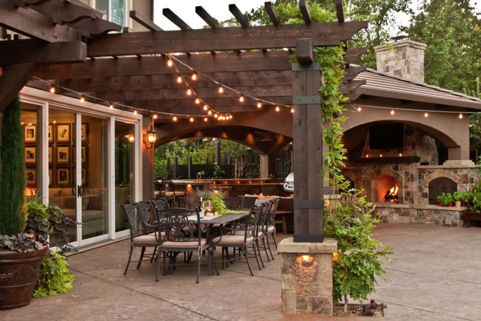 Outdoor patio with dining table, pergola, and stone fireplace. String lights, greenery, and open air bar.