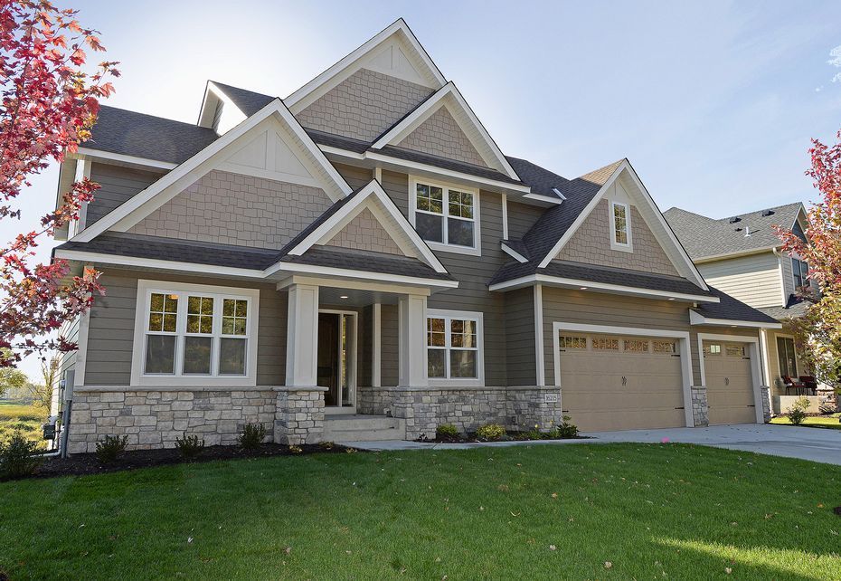 Two-story house with tan siding, stone base, and a green lawn. Brown garage doors and a blue sky.