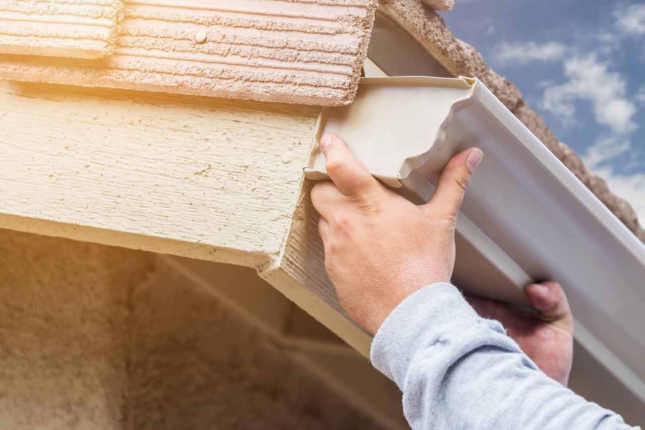 Hands installing white gutter on a home, under beige eaves. Blue sky in background.