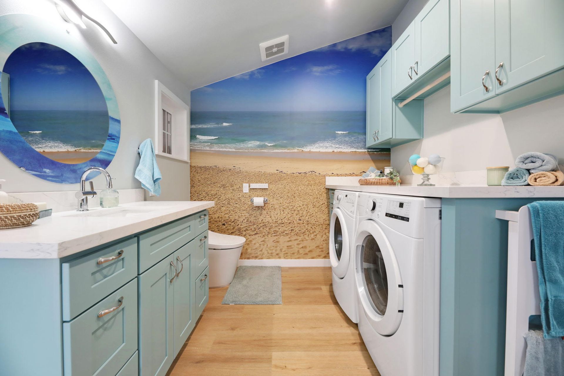 Bathroom/laundry room combo with beach scene mural, blue cabinets, washing machines, toilet, and sink.