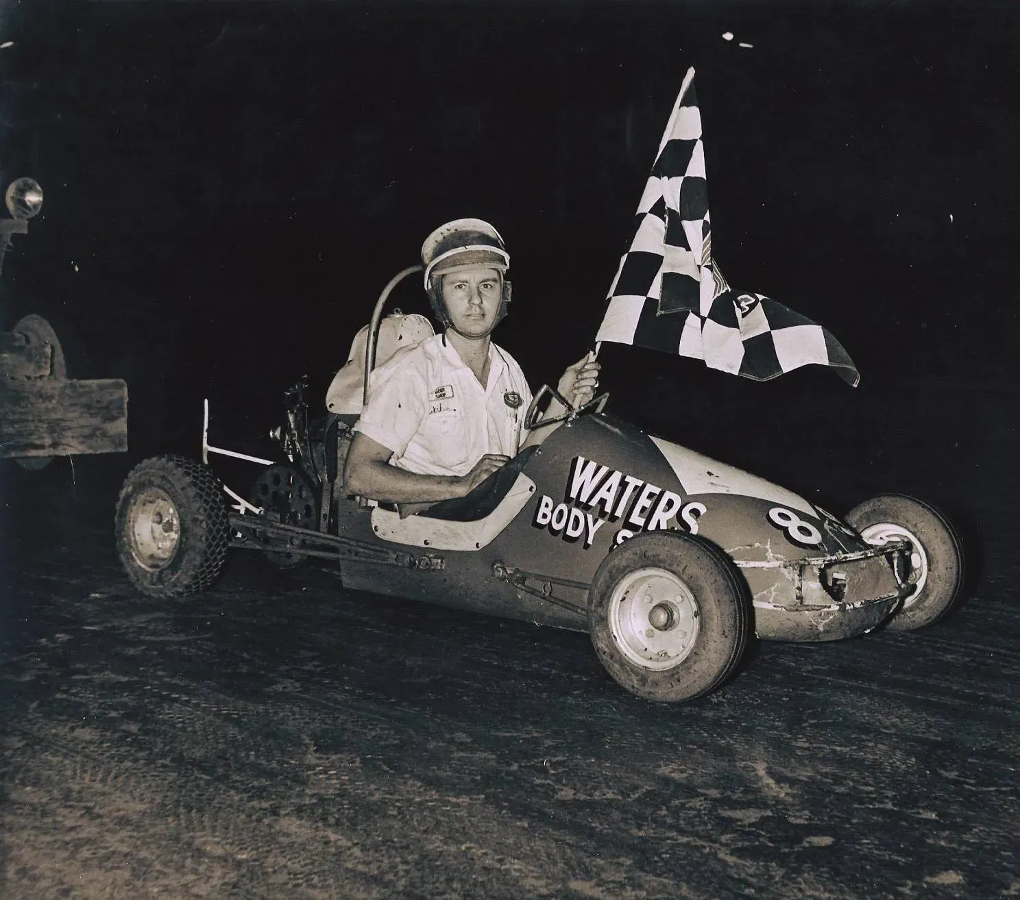 A racer drives a small go-kart, holding a checkered flag on a dirt track.