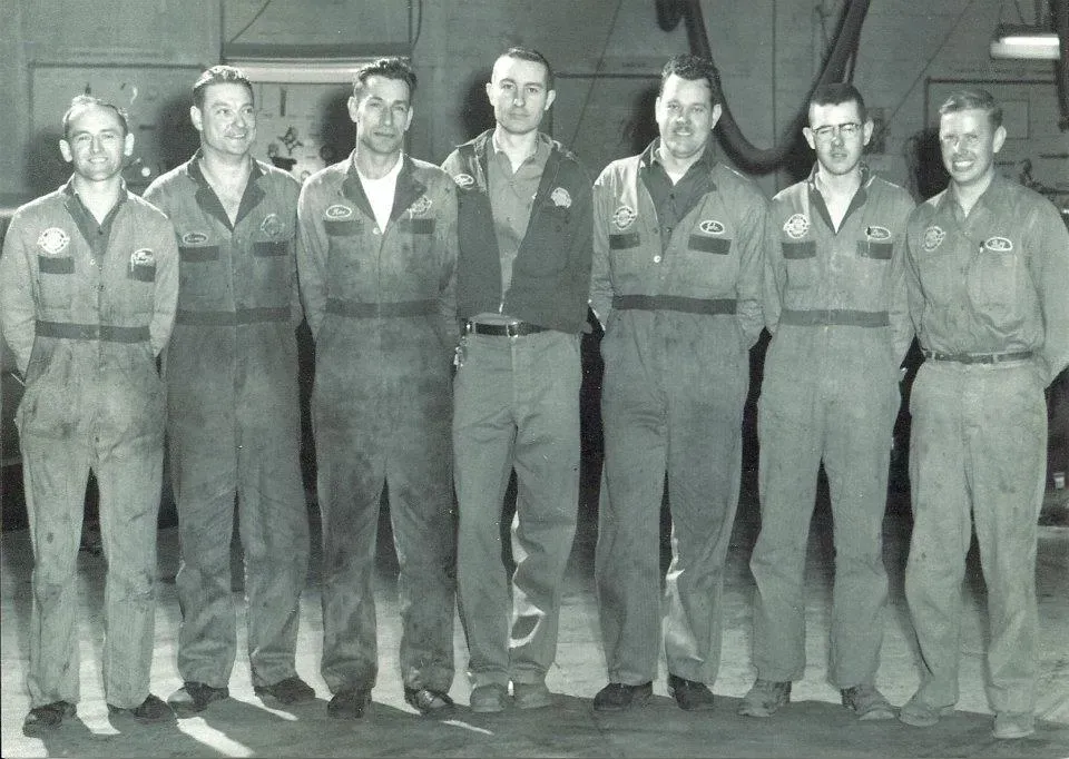 Seven uniformed workers stand in a row inside a hangar, facing the camera.