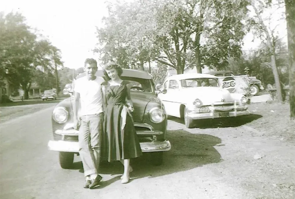 Two people standing by a vintage car with its hood open on a tree-lined street