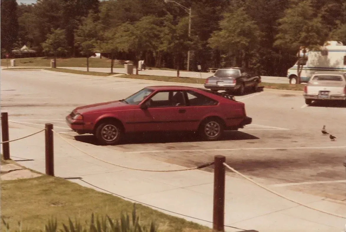 Red car parked in a nearly empty lot with a few other cars and trees in the background.