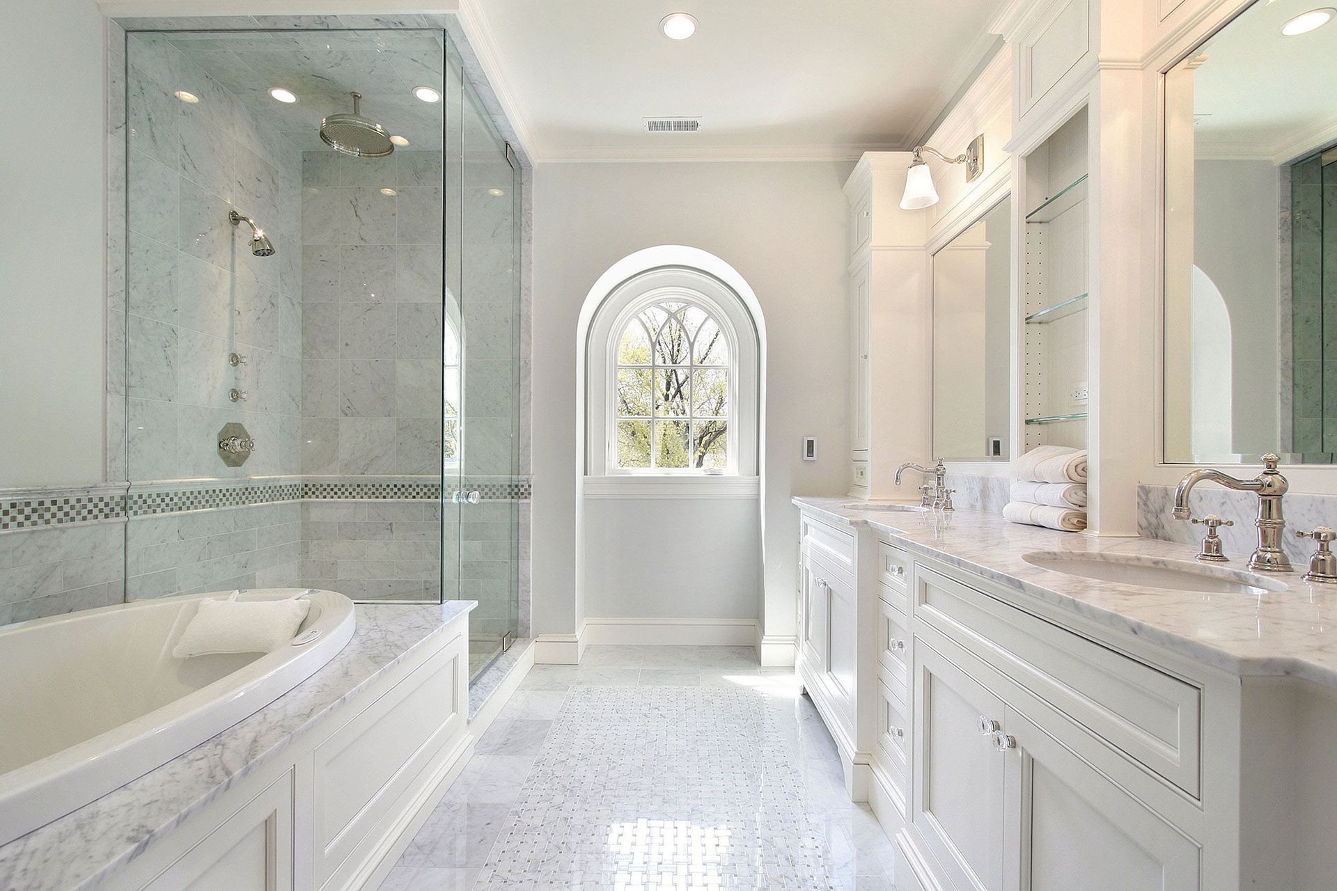Elegant white bathroom with glass shower, tub, and vanity; arched window.