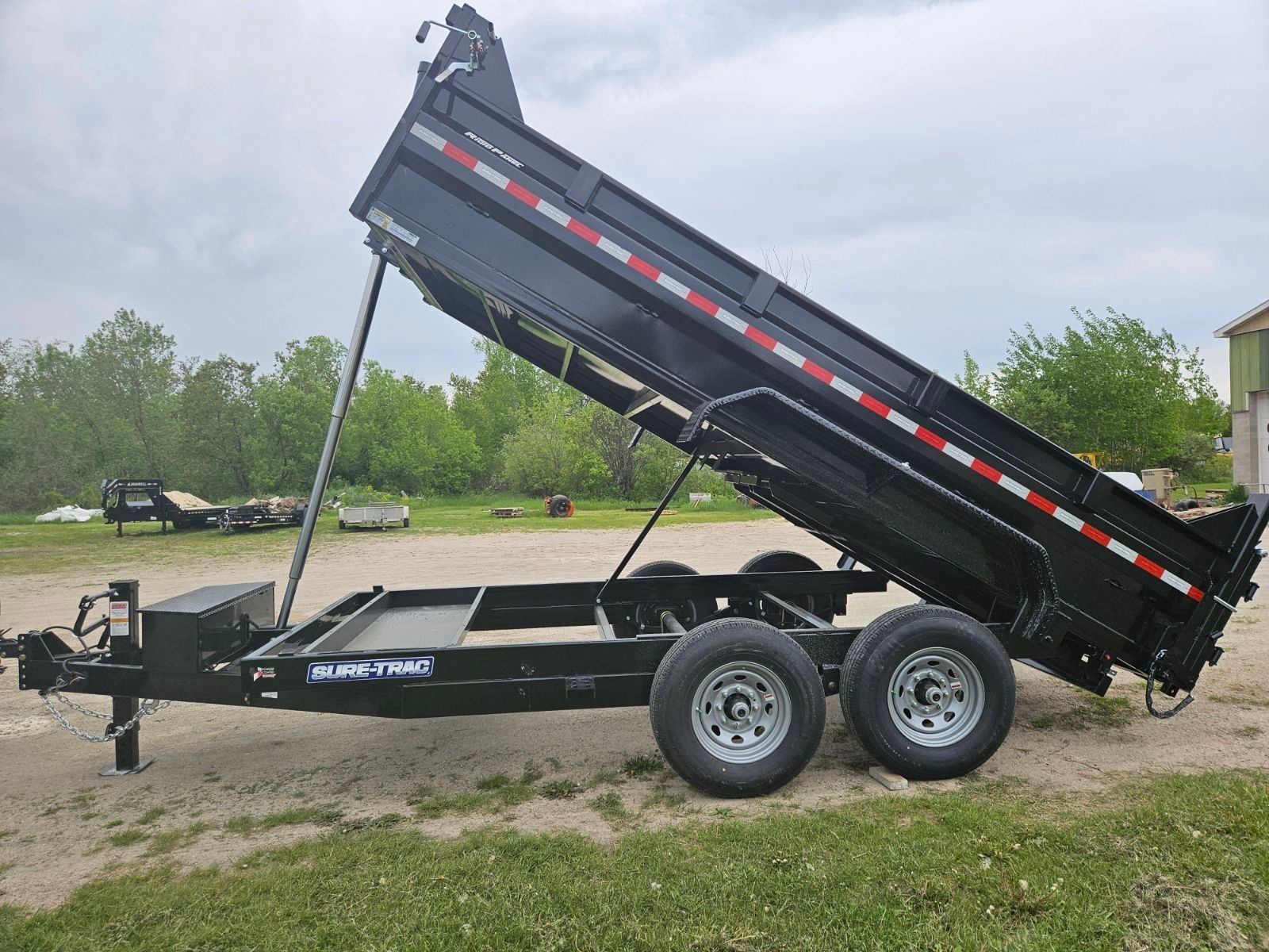 Black dump trailer tipped to unload, with raised bed and tandem wheels on a dirt lot