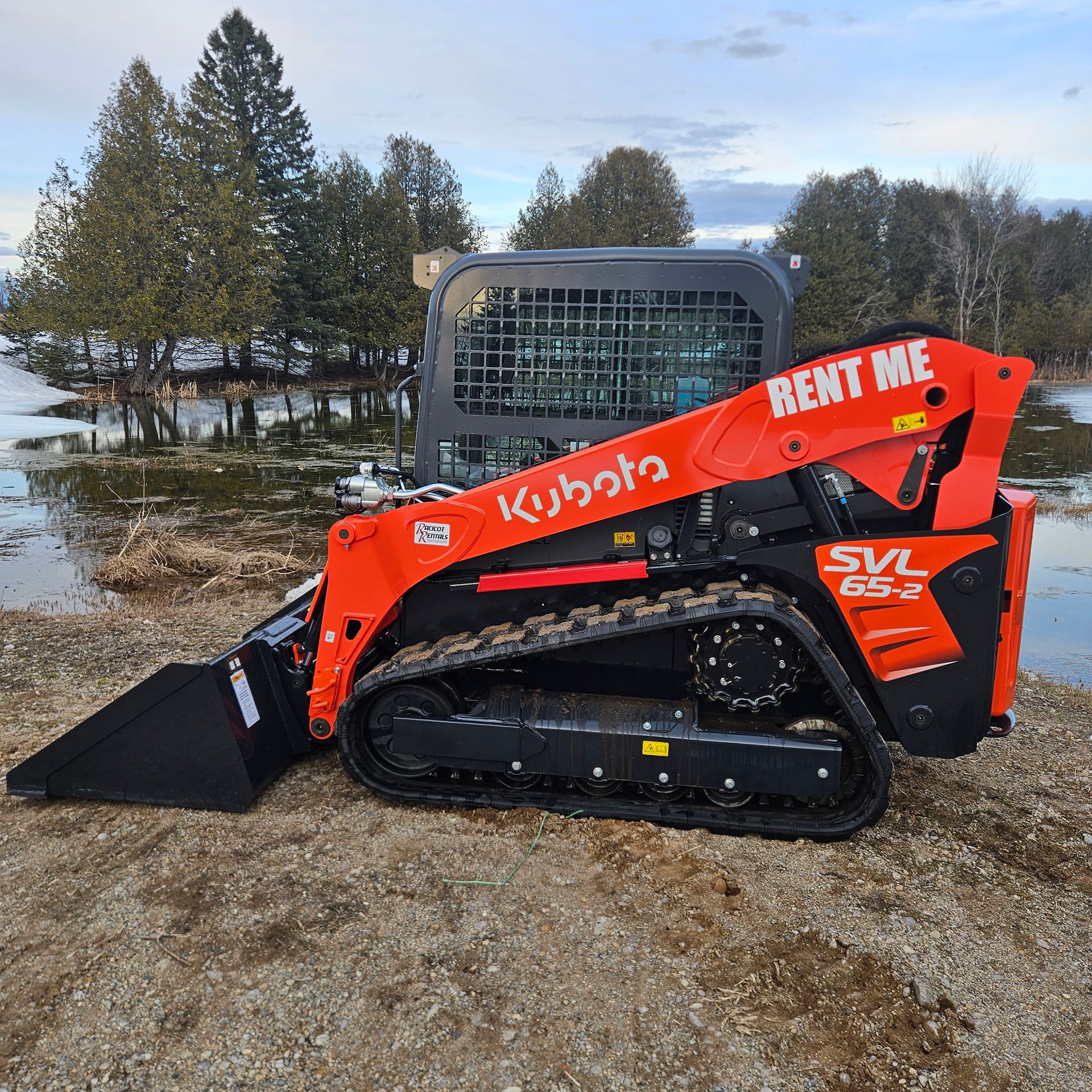 Orange Kubota skid steer with bucket in muddy flooded area near trees and a trailer.
