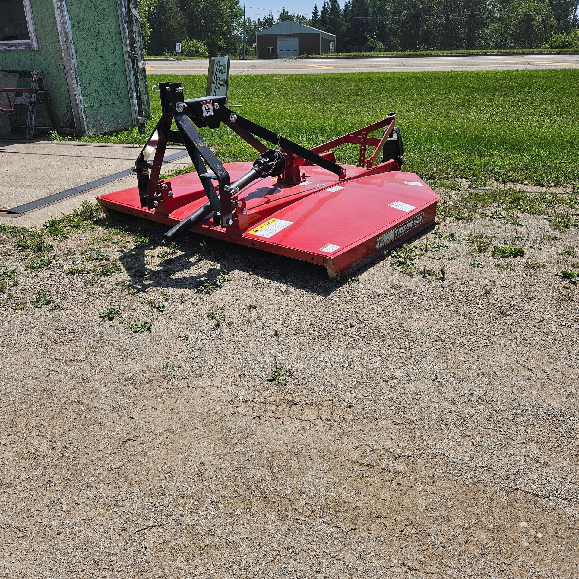 Red mower deck attachment on gravel beside a green building, with grass field and trees in the background