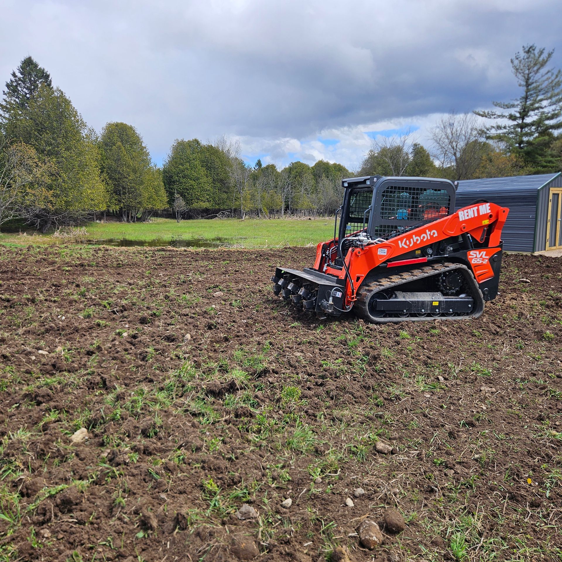 Orange compact track loader on a muddy field near trees and a shed under cloudy skies