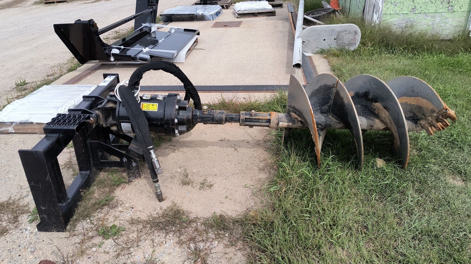 Black tractor attachment with curved auger blades on a driveway beside grass and a wall