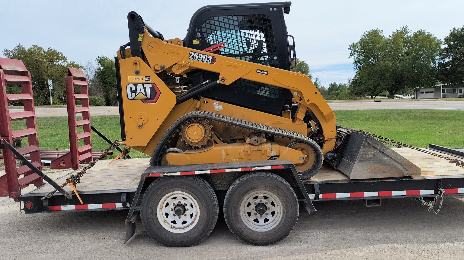 Yellow CAT skid steer on a flatbed trailer, parked beside a road in a grassy field.
