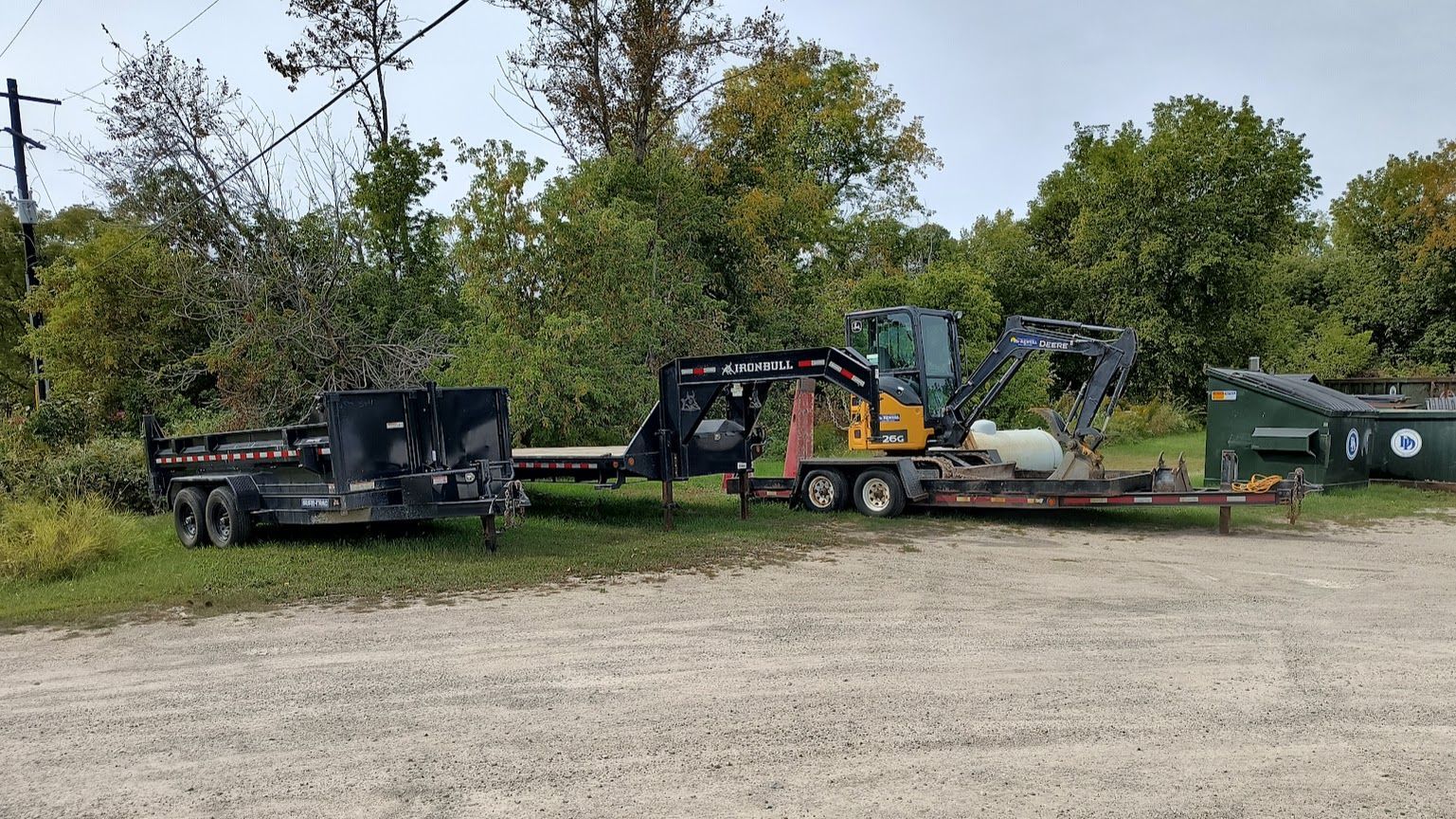 Construction equipment parked on gravel beside trees and a green tank in a rural lot