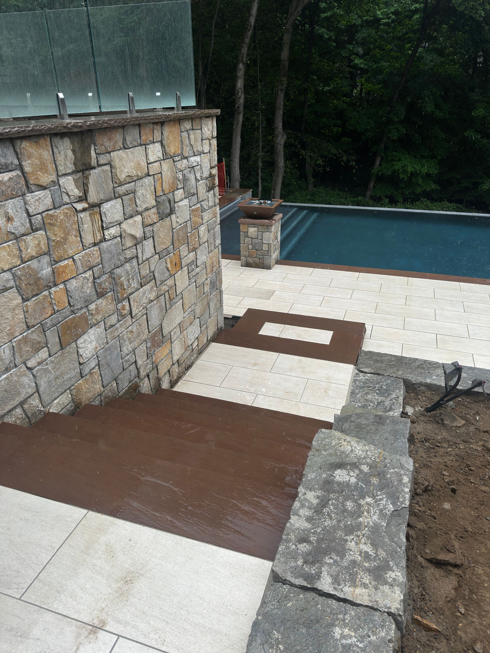 Stone patio with steps, a retaining wall, and a swimming pool beside a wooded area