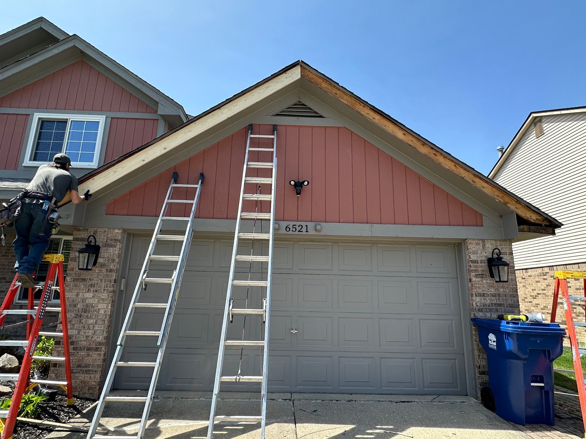 Man on a ladder working on the roof trim above a garage door. Ladders, house numbers, and a blue trash can are visible.