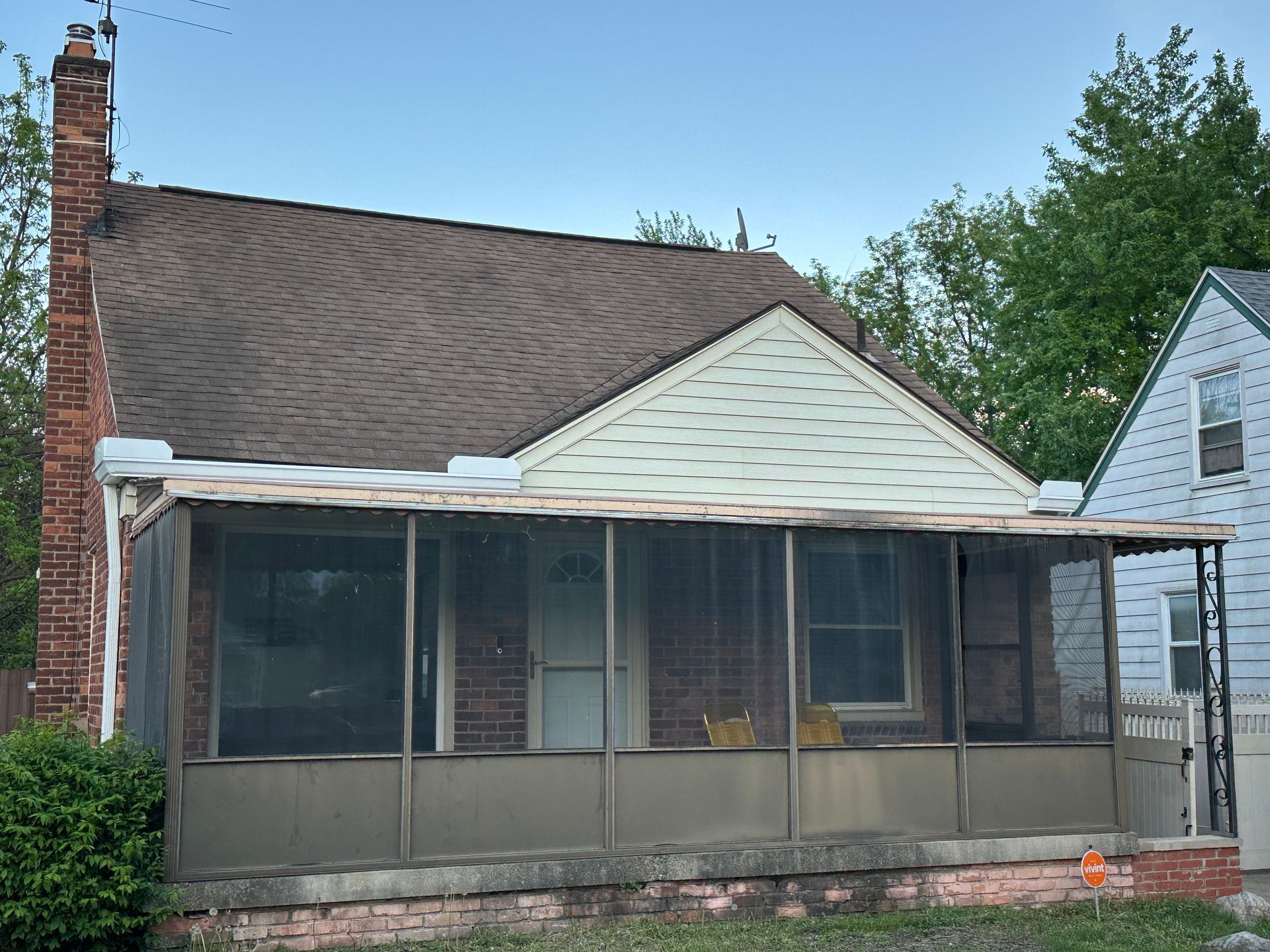 Low-angle view of a small brick house with a screened-in porch and chimney. Brown roof, beige siding.