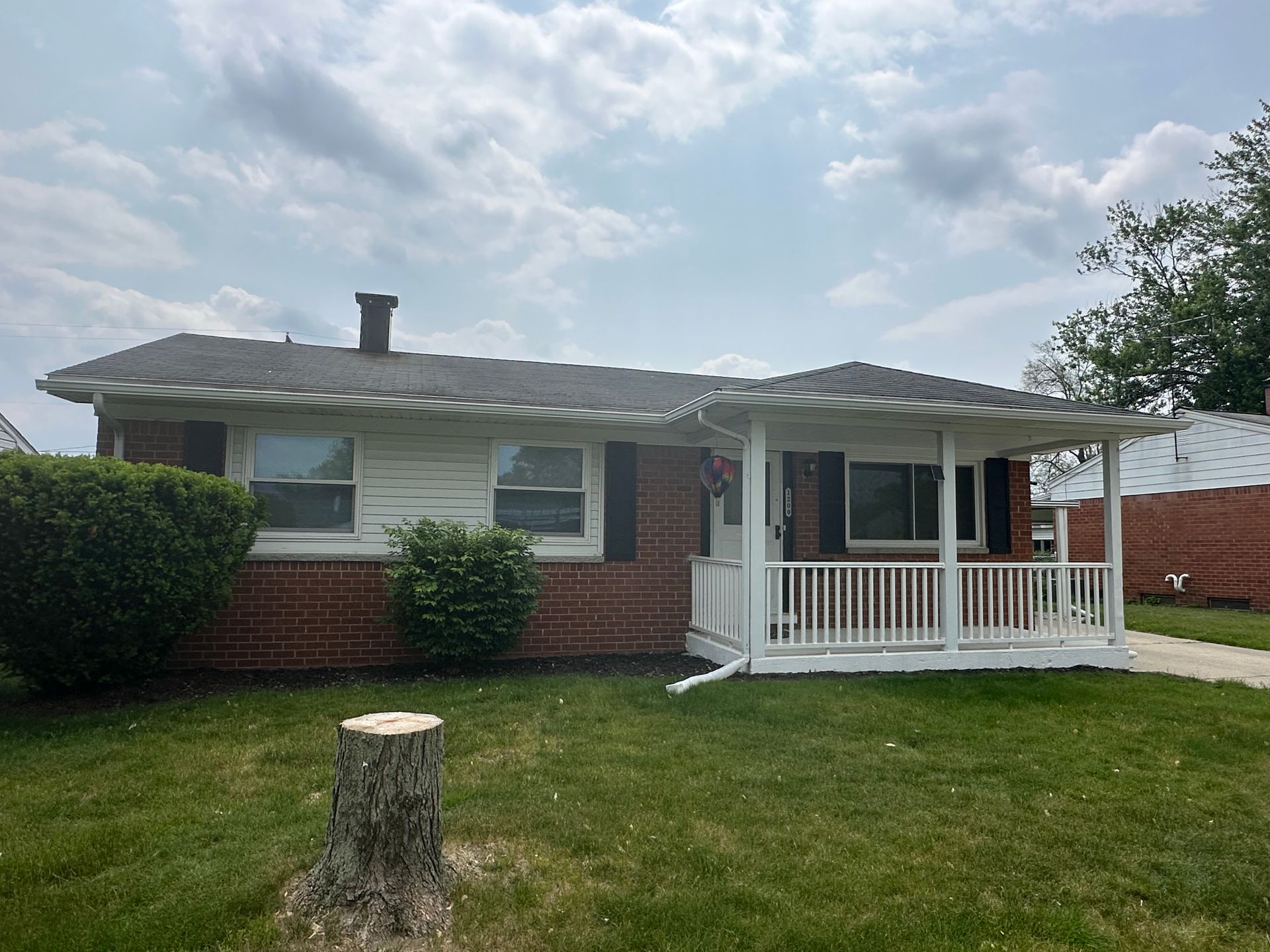 Brick ranch house with small porch, black shutters, and green lawn under a cloudy sky.
