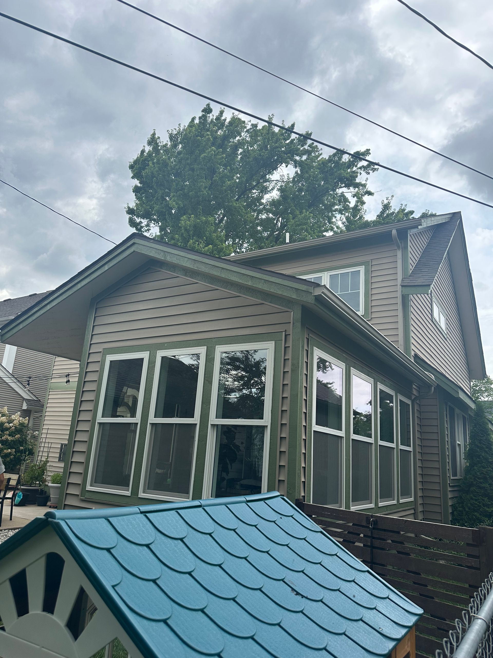 Tan and green house with many windows, cloudy sky, blue playhouse in foreground.