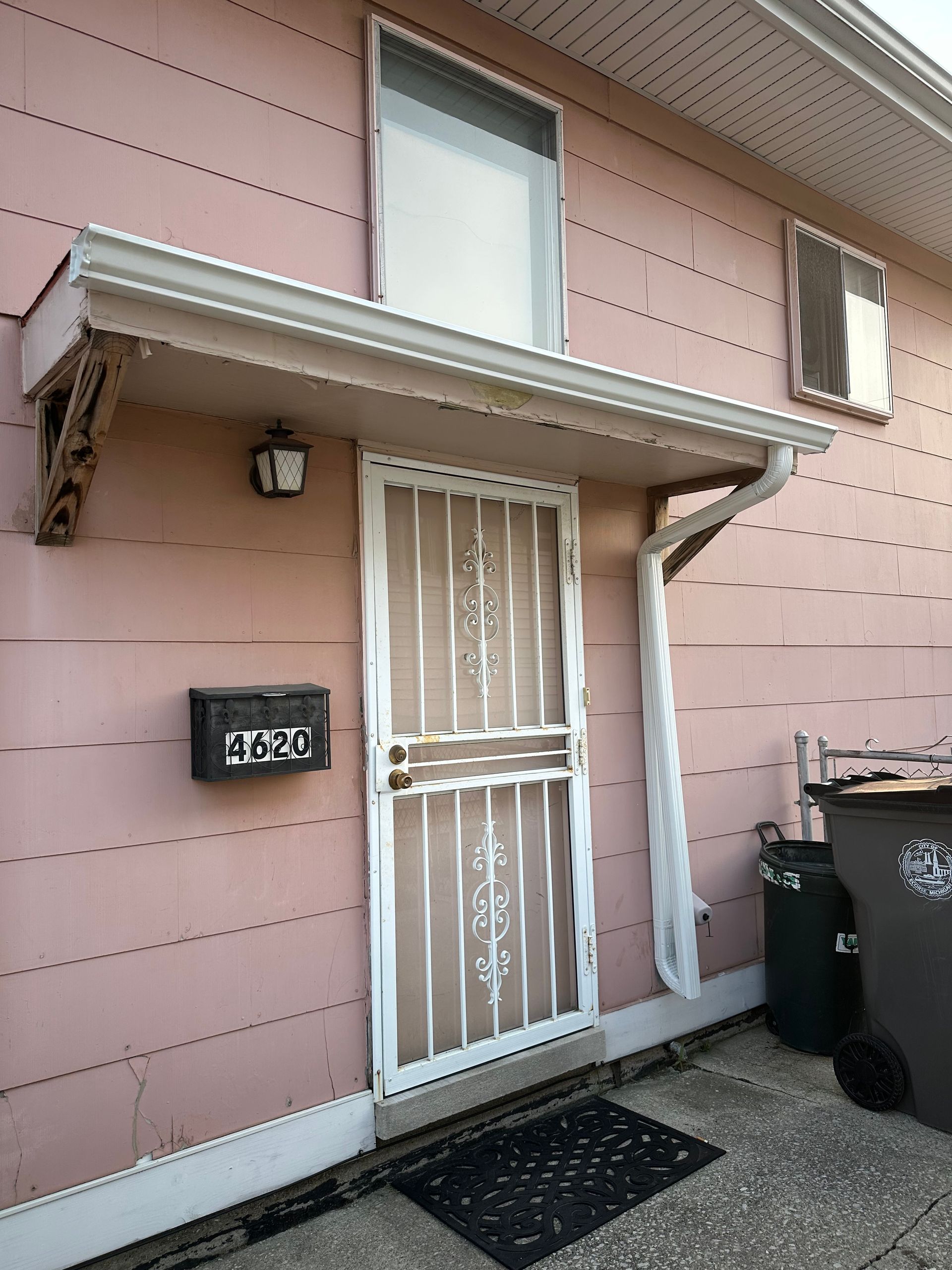 Pink house exterior with security door and awning. Black mailbox, trash cans on the right.
