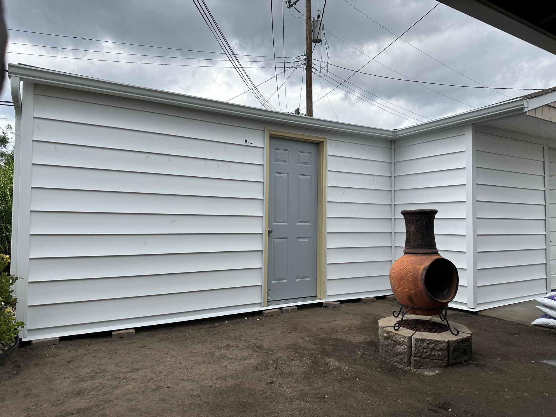 White-sided shed with a gray door, next to a brick fire pit, under a cloudy sky.
