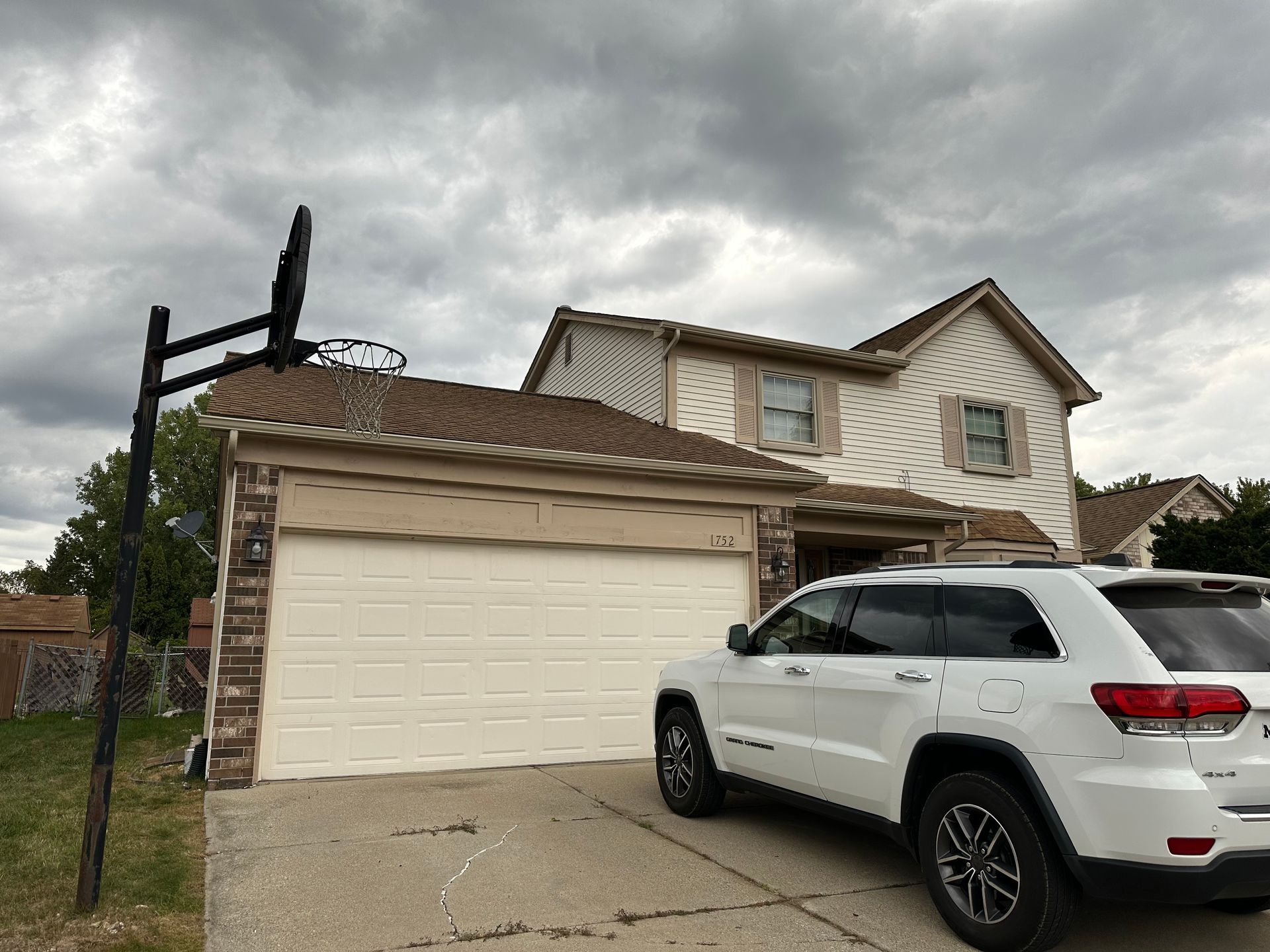 White SUV parked in front of a beige two-story house with a basketball hoop on a cloudy day.
