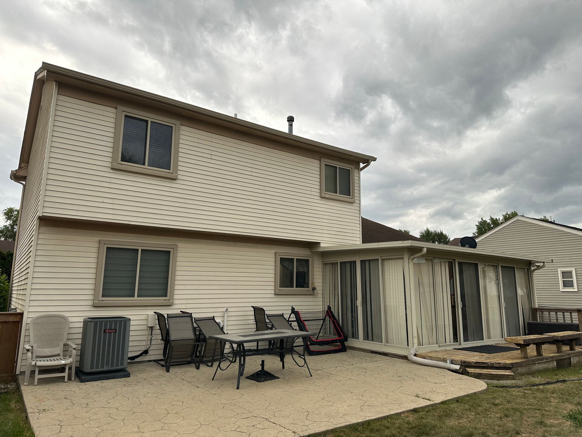 Back of a two-story beige house with a concrete patio. A sunroom and patio furniture are visible. Overcast sky.