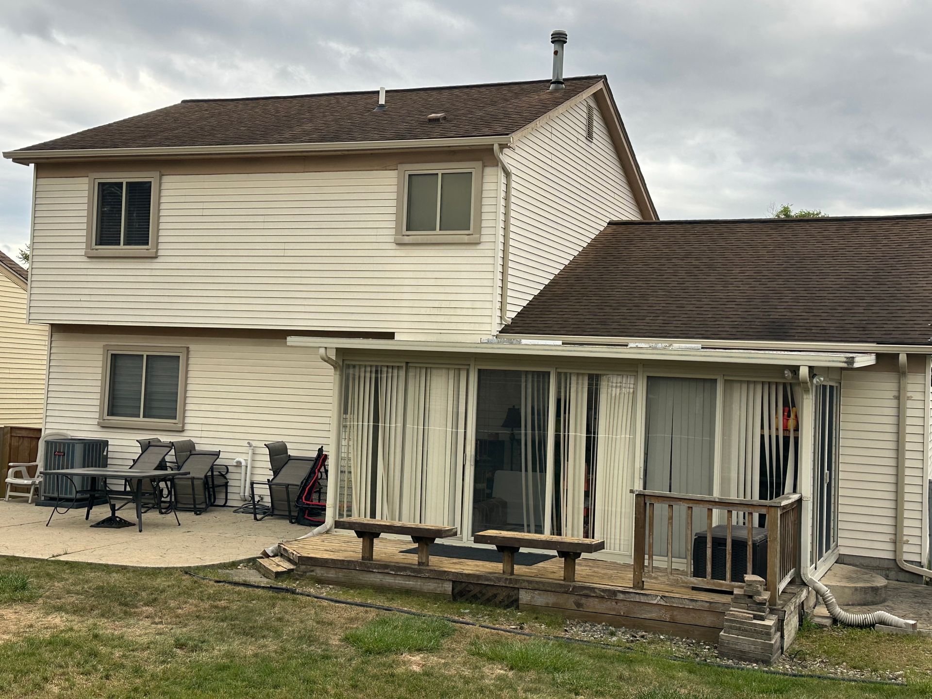 Two-story house with a patio and deck; beige siding and brown roof. Curtains hang from the deck roof.