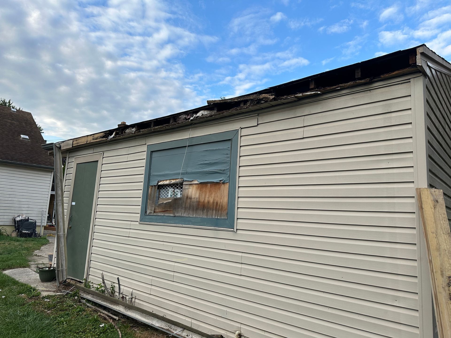 Exterior of a damaged beige shed with a crumbling roof, a weathered window, and a green door against a cloudy sky.