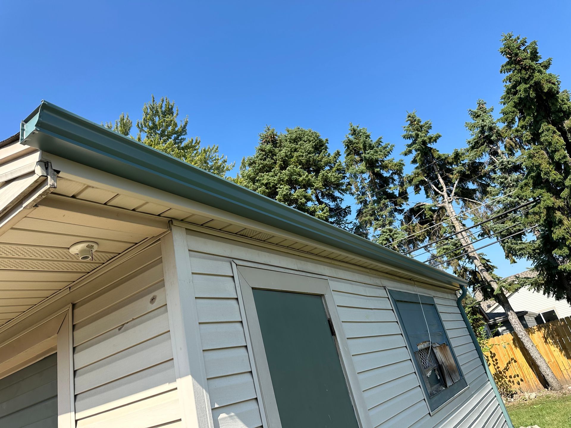 Small building with light-colored siding, green trim, and a blue sky with trees in the background.