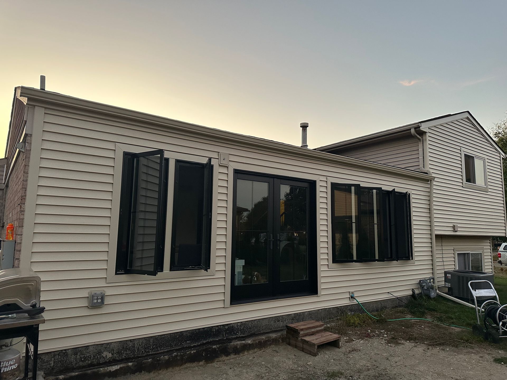 Rear view of a light beige siding house with black shutters, doors, and window frames against a dusk sky.