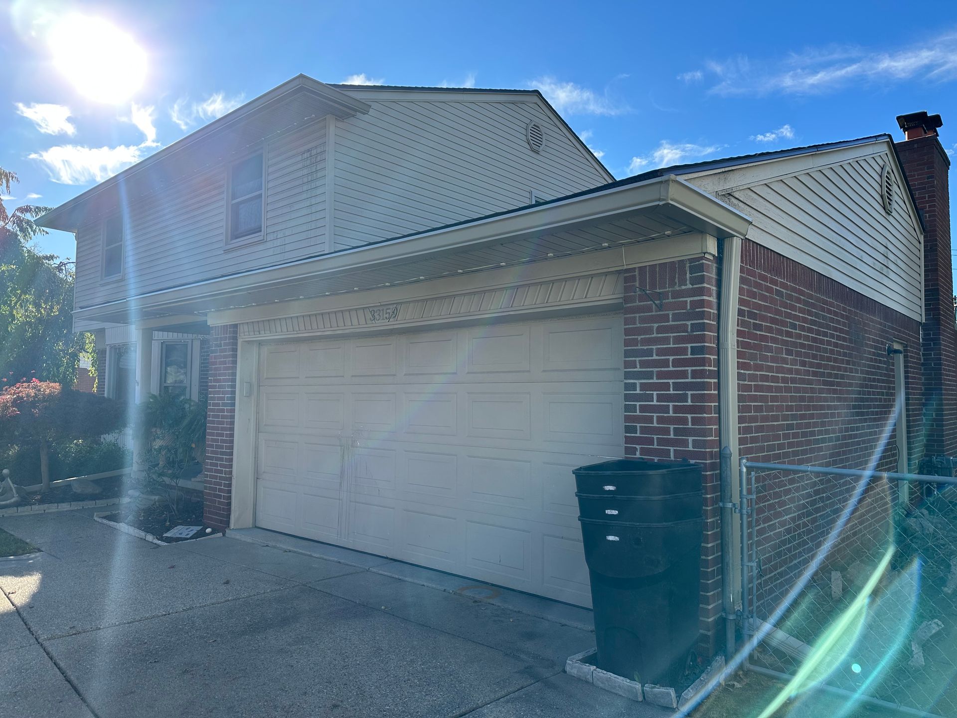 Two-story house with beige garage doors and a brick exterior under a blue sky. A black trash can sits near the garage.