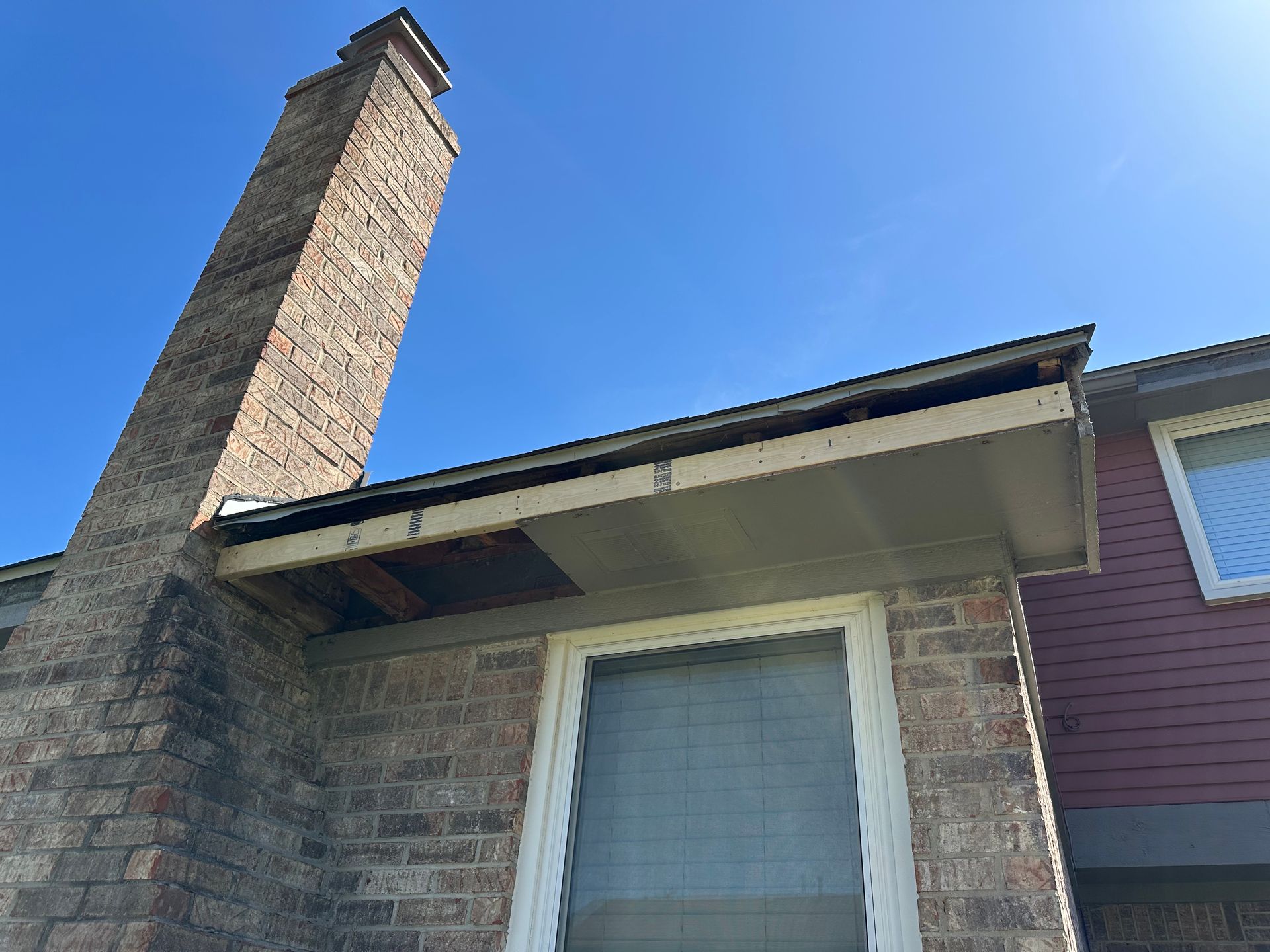 Brick chimney next to a roof with damaged wood and a clear blue sky above.