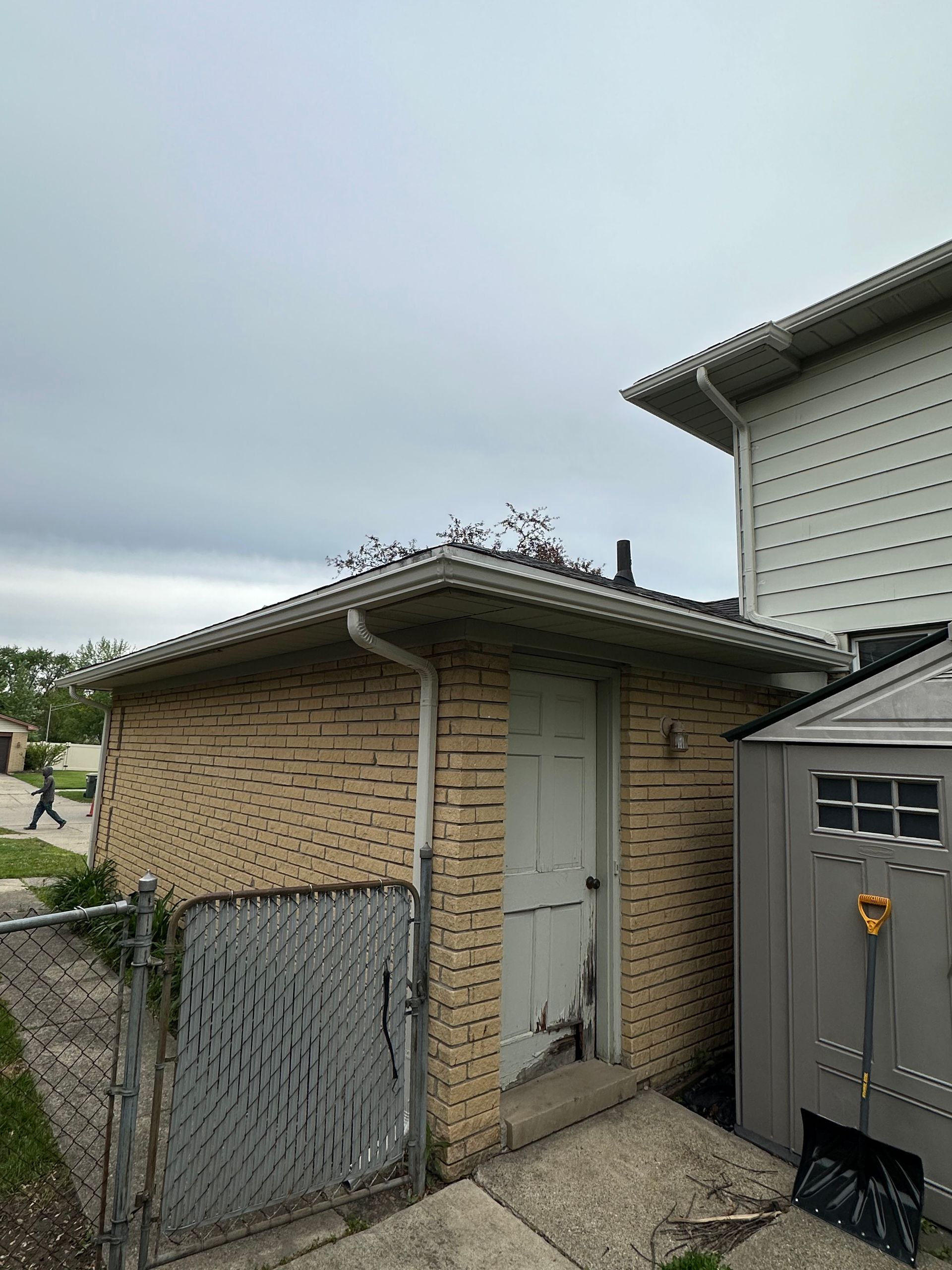 Brick shed with a white door, a chain-link fence, and a gray storage unit, under a cloudy sky.