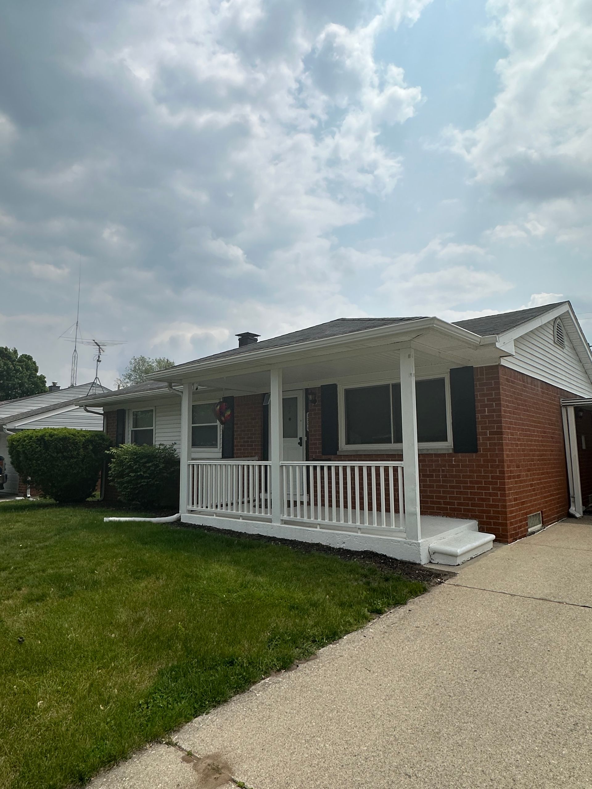 Brick ranch house with white porch, dark shutters, green lawn, and cloudy sky.