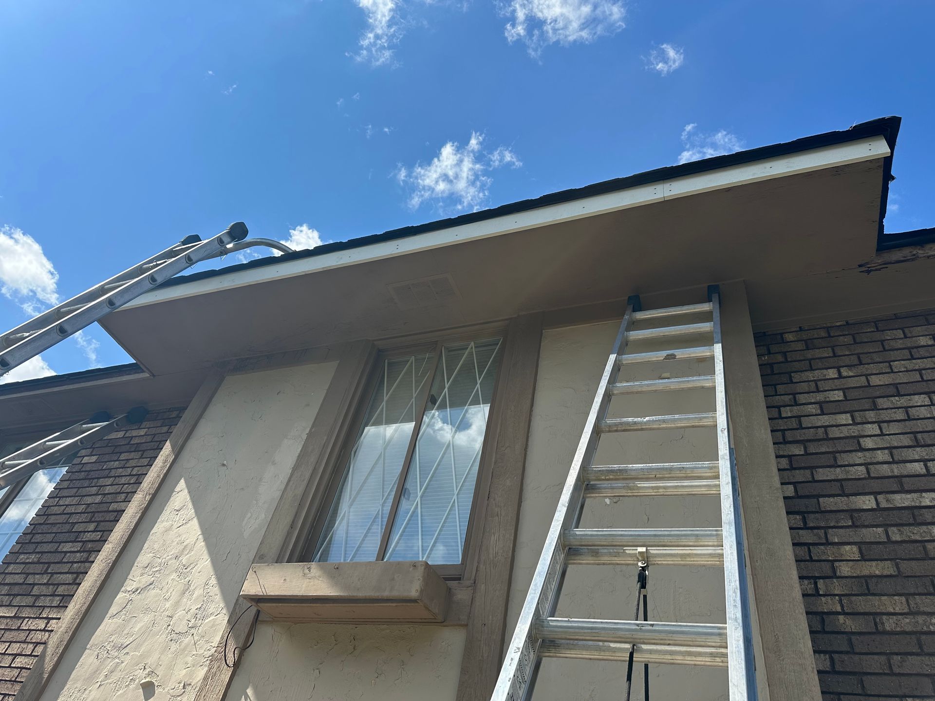 Ladders against a building exterior with brown trim, beige stucco, and brick, under a partly cloudy blue sky.
