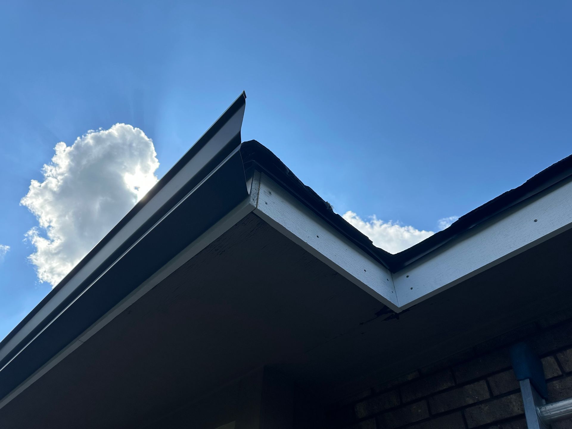 Low-angle view of a roof corner with black trim, white eaves, and a blue sky with clouds.
