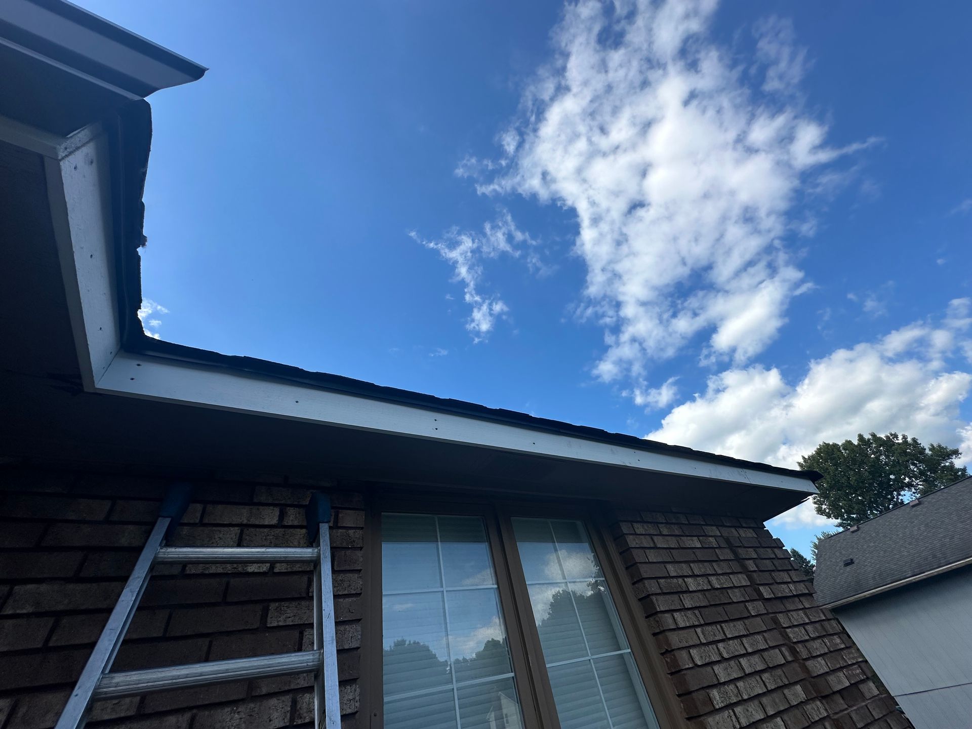 Ladder leaning against a house with a damaged roof, set against a blue sky with clouds.