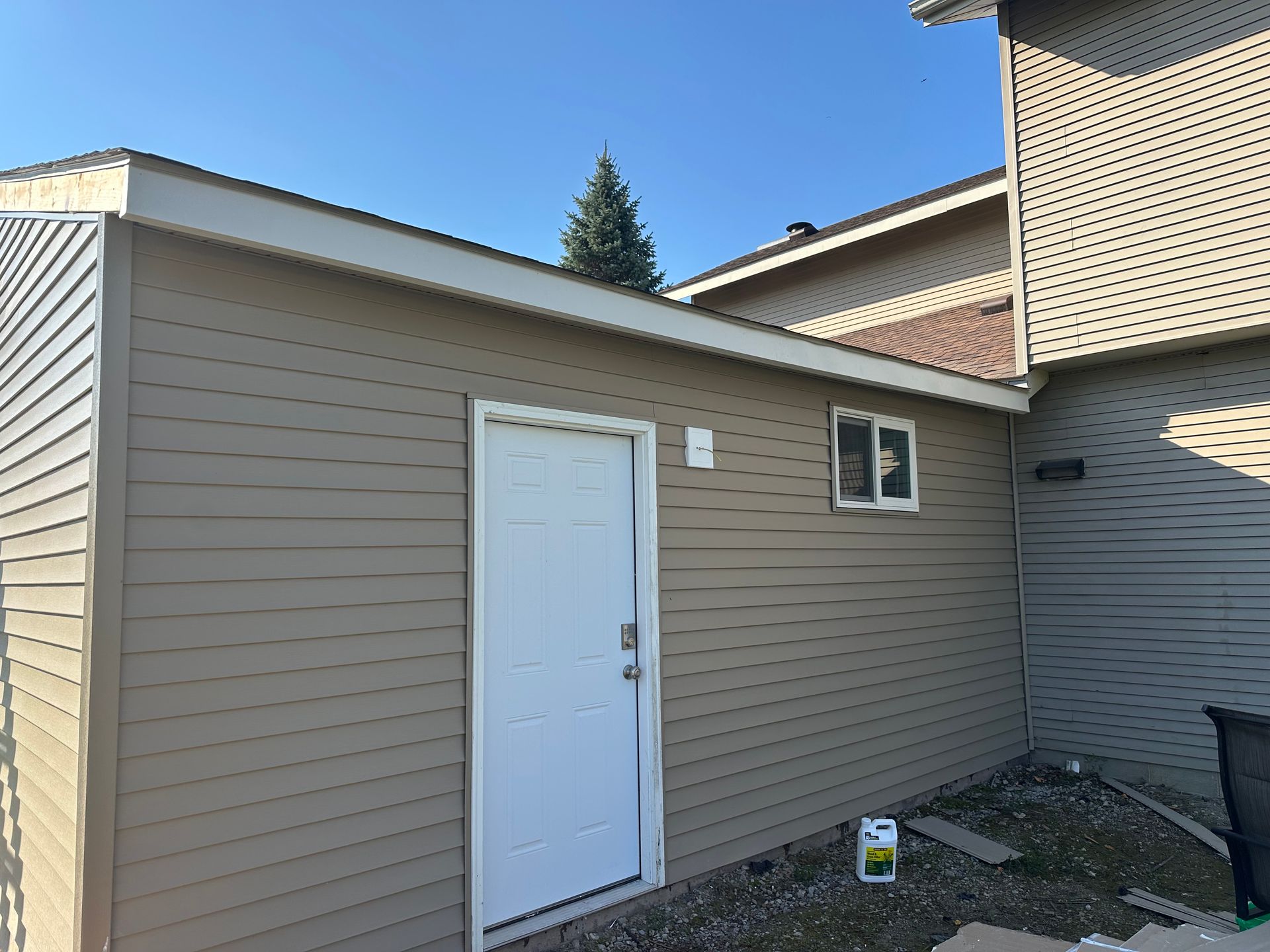 Tan-sided shed with white door, small window, and light-colored trim against a clear blue sky.
